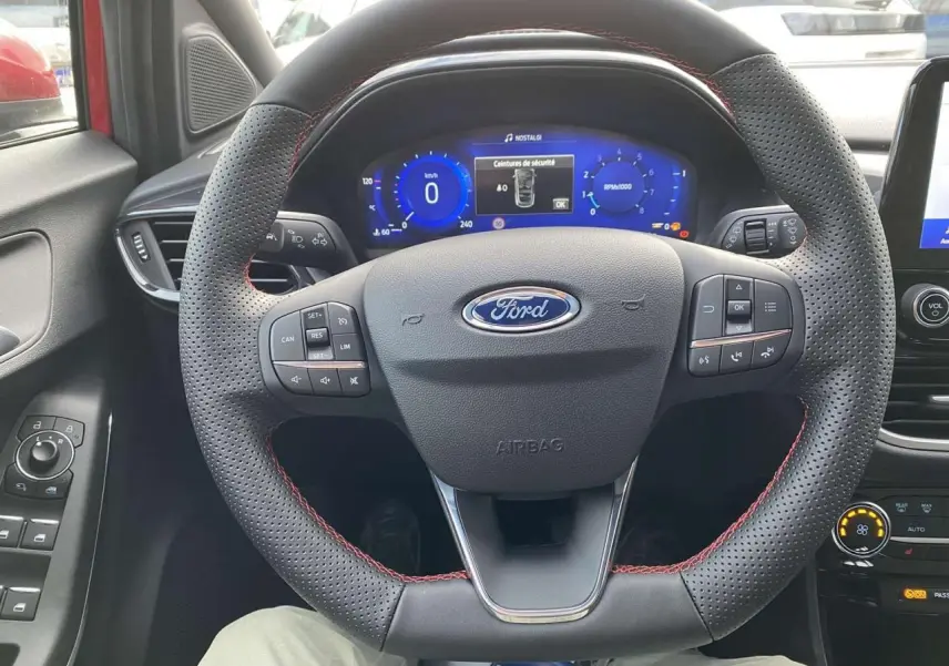 Vue intérieure centrée sur le volant perforé avec surpiqûres rouges du Ford Puma 2022, tableau de bord numérique allumé.