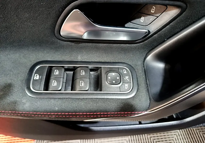 Gros plan sur la commande des vitres et rétroviseurs côté conducteur d'une Mercedes Classe A noire, avec surpiqûres rouges.