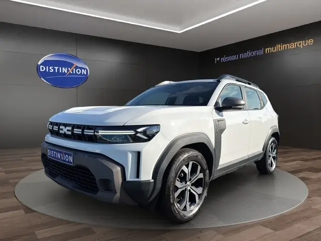 Dacia Duster blanc en 3/4 avant droit, avec toit noir, jantes alliage et calandre moderne, sur podium intérieur showroom.