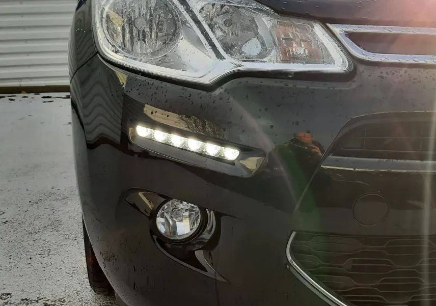 Gros plan sur l'avant gauche noir d'une Citroën C3 2016 avec feux diurnes LED allumés et carrosserie mouillée.