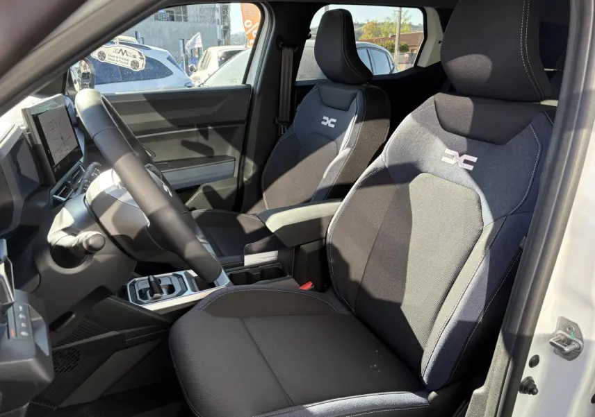 Vue intérieure côté conducteur du Dacia Duster blanc métallisé 2024, sièges Denim avec logo réfléchissant et console centrale.