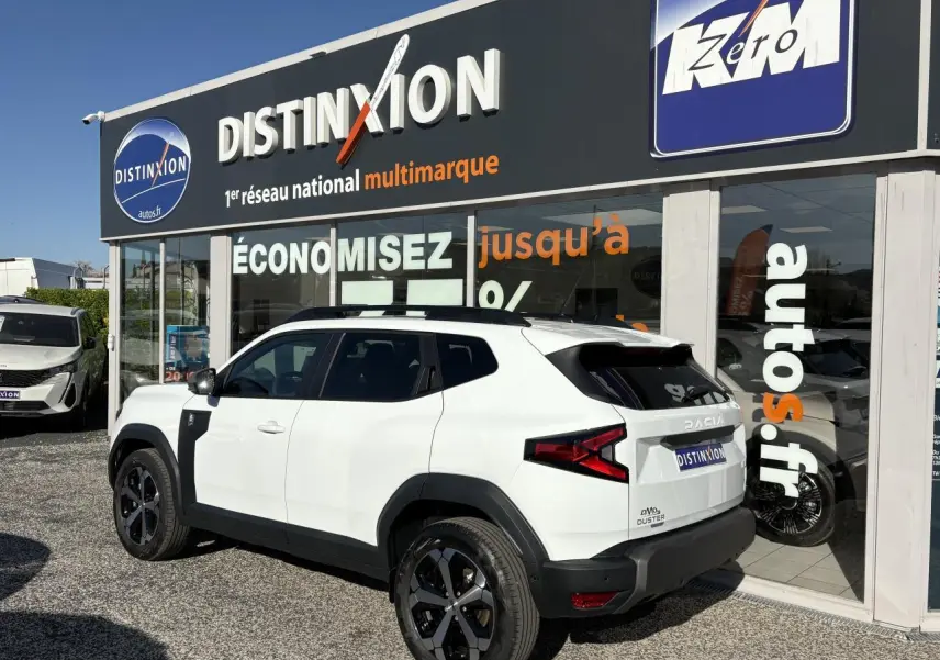 Vue 3/4 arrière droite d’un Dacia Duster blanc métallisé avec barres de toit noires et jantes alliage 18 pouces.