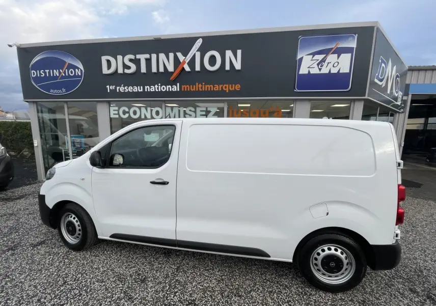 Vue de profil côté gauche d’un utilitaire Opel Vivaro blanc stationné devant un concessionnaire.