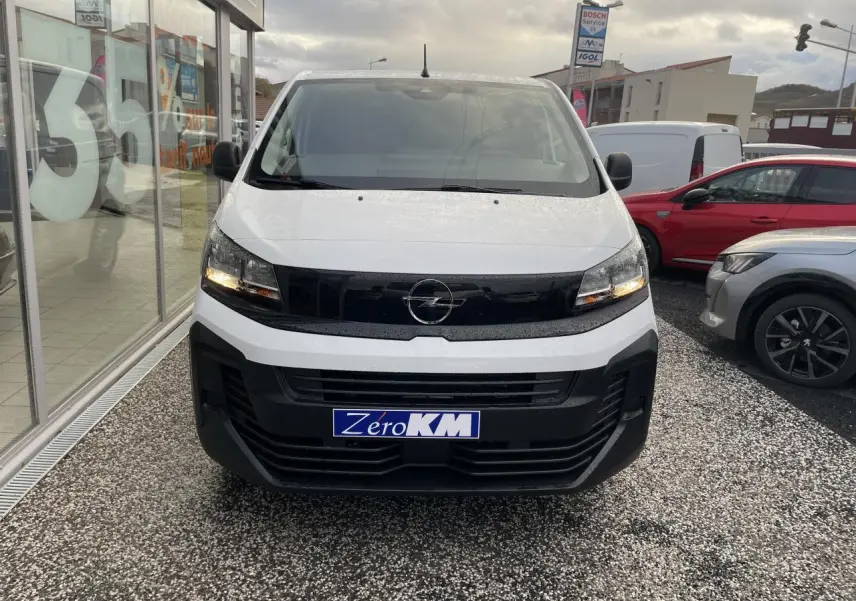 Vue de face d’un Opel Vivaro blanc 2024 avec calandre noire et feux allumés sur parking.