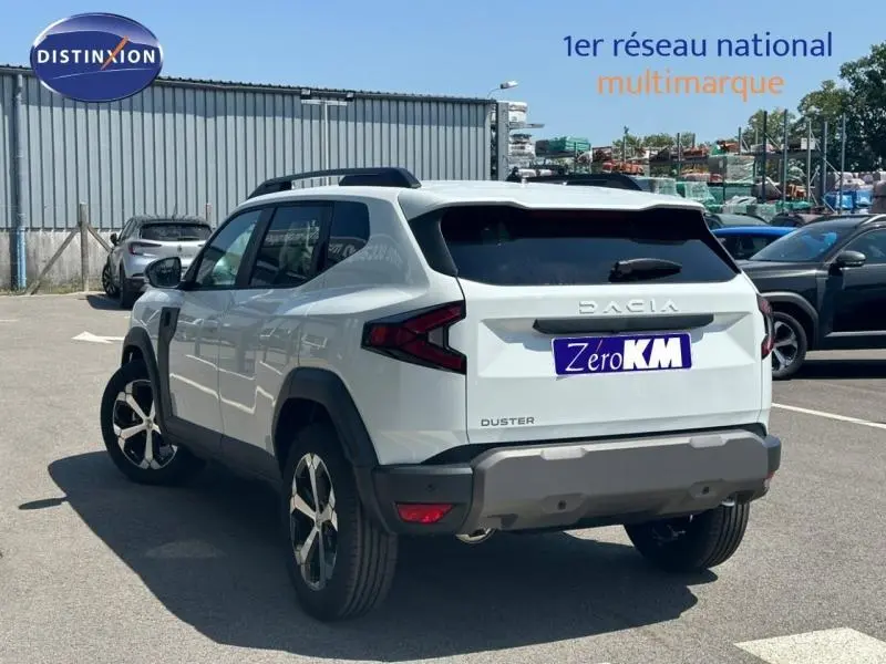 Vue 3/4 arrière droite d'un Dacia Duster blanc Glacier avec barres de toit et jantes alu, stationné en extérieur.