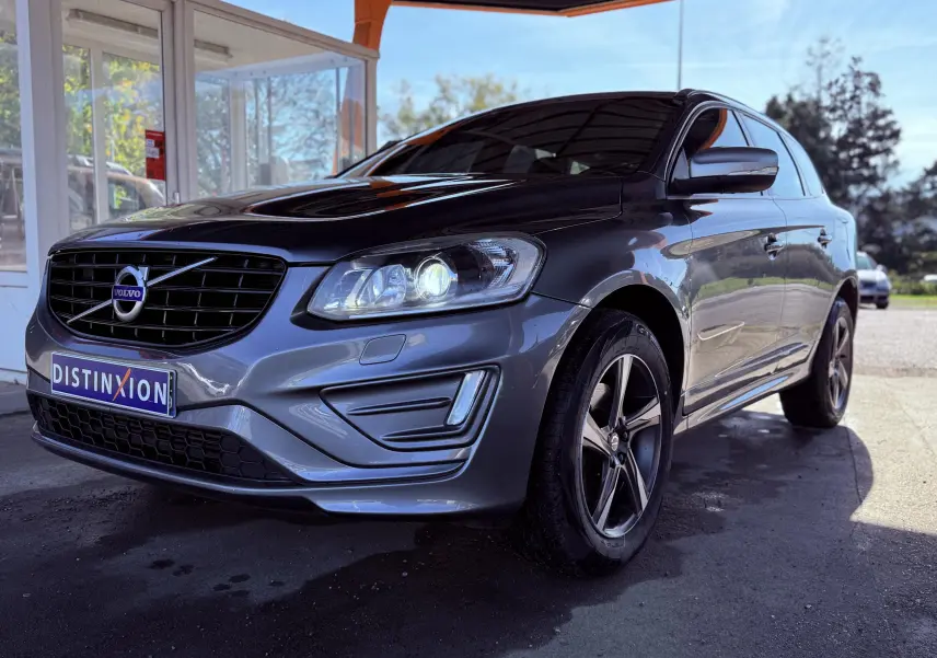 Volvo XC60 gris en 3/4 avant droit, phare allumé et jantes alliage cinq branches visibles, stationné devant un bâtiment.