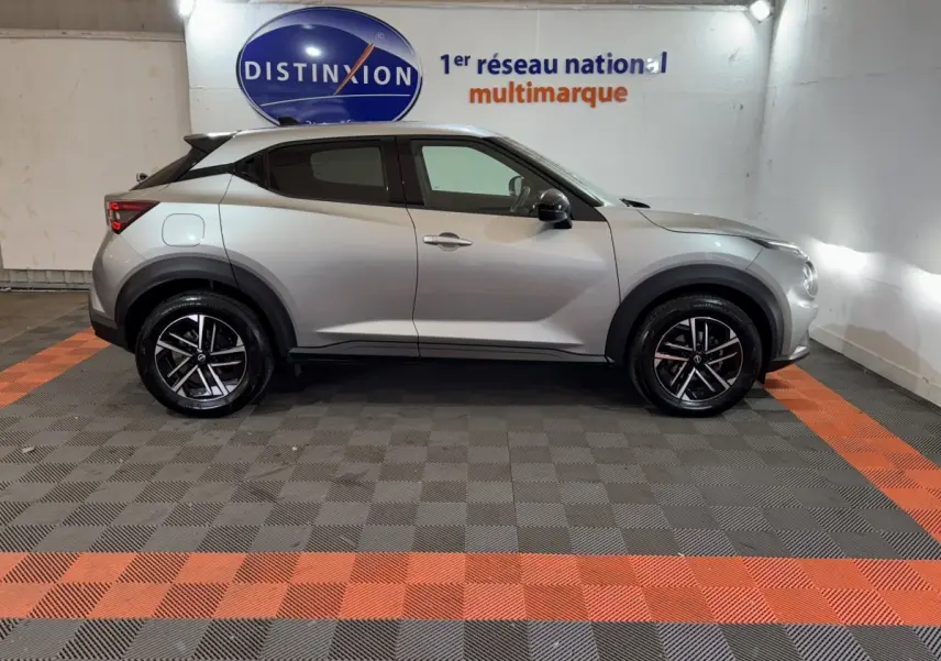 Profil droit d’un Nissan Juke gris clair 2025, avec jantes noires et détails noirs contrastants, en intérieur showroom.