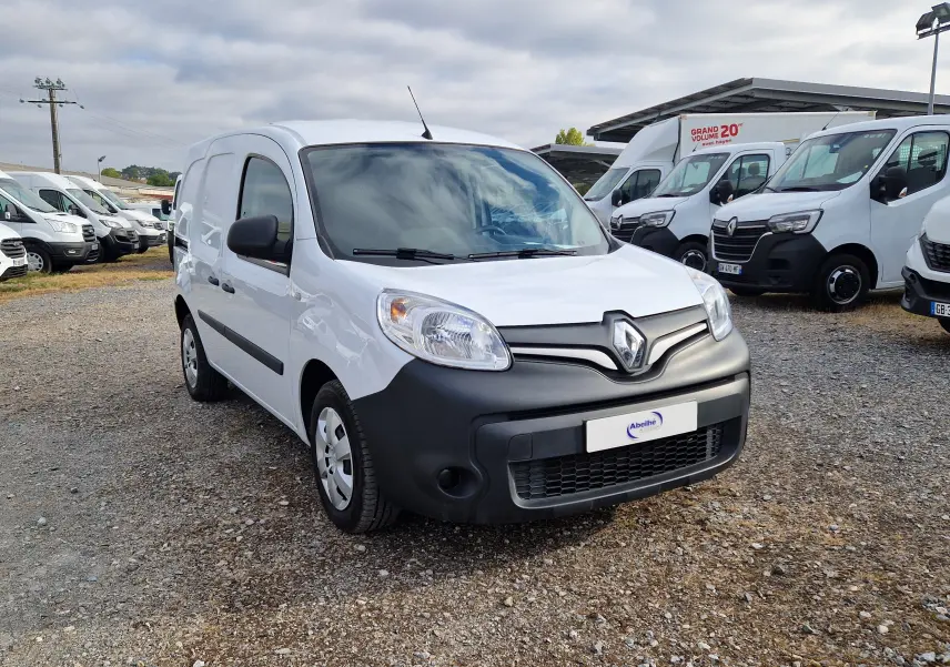 Vue 3/4 avant d’un Renault Kangoo blanc avec pare-chocs noir mat, stationné sur un sol gravillonné en extérieur.