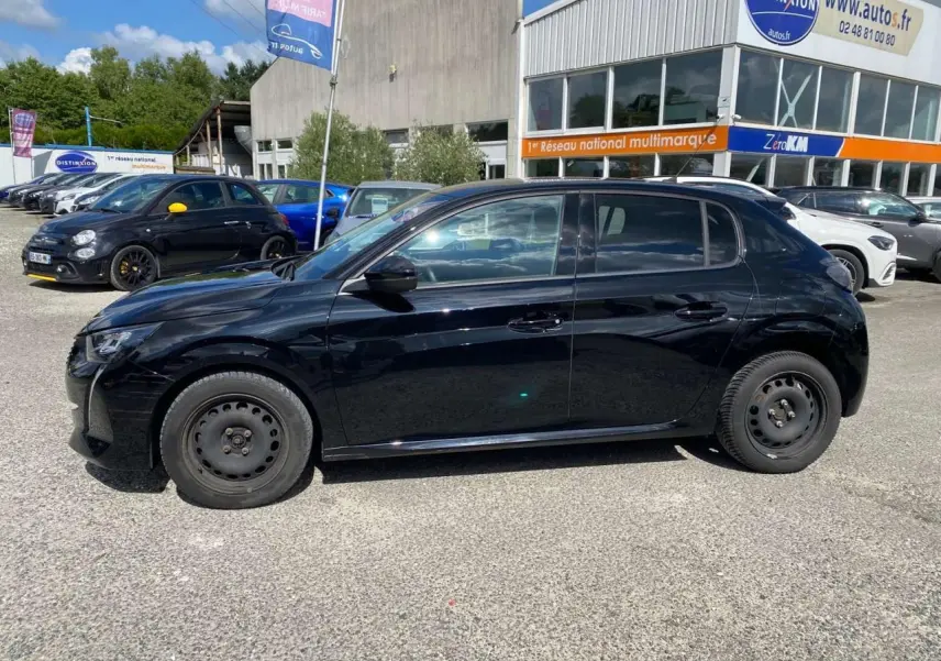 Peugeot 208 noire vue de profil côté gauche, stationnée sur parking devant un bâtiment commercial.