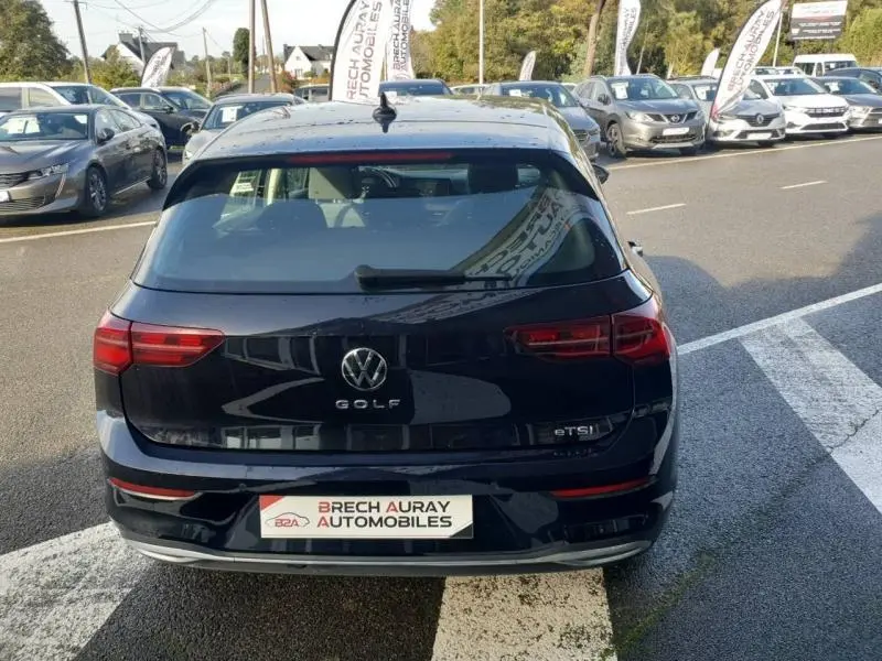 Vue arrière d'une Volkswagen Golf noire intense nacrée garée sur un parking avec plusieurs autres voitures visibles en arrière-plan.