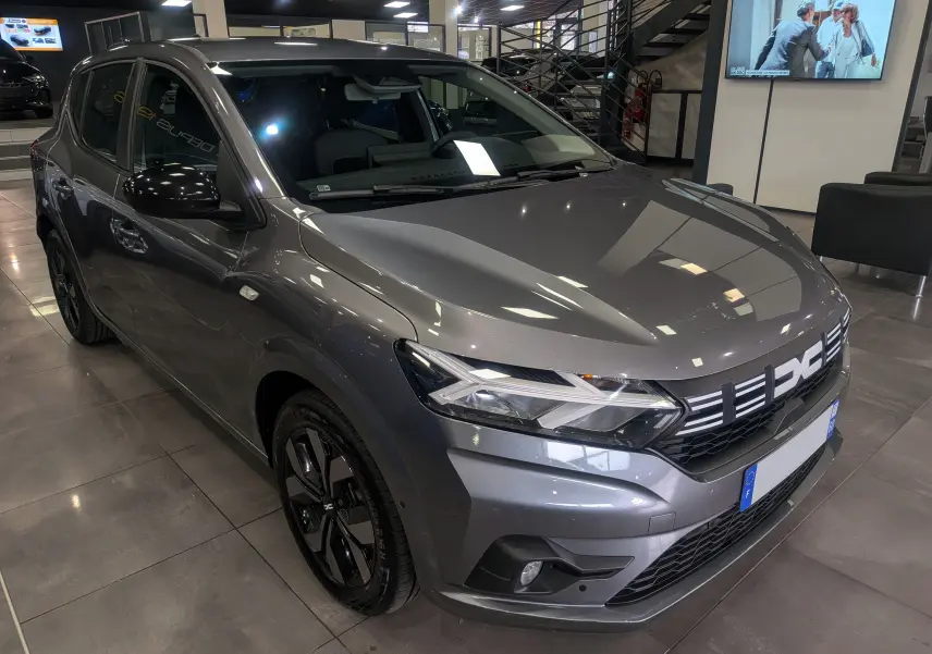 Vue 3/4 avant d'une Dacia Sandero TCe 90 CH Journey gris schiste avec jantes noires et calandre lumineuse en showroom.
