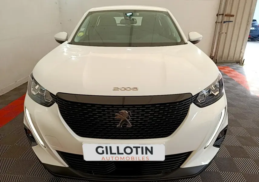 Vue avant d'une Peugeot 2008 blanche avec calandre noire et phares LED, stationnée en intérieur sur sol damier.