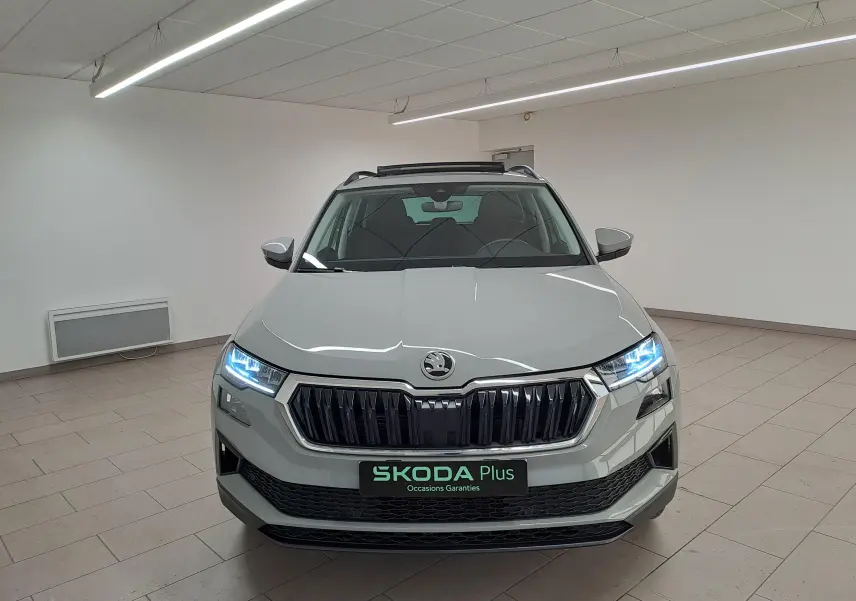 Vue frontale d'un Skoda Karoq gris perle 2023 avec phares allumés et toit ouvrant panoramique visible en intérieur.