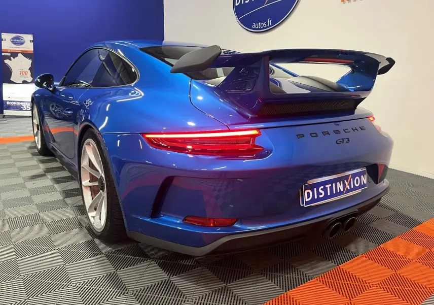 Vue 3/4 arrière droite d'une Porsche 911 GT3 bleu saphir avec aileron arrière et feux LED allumés en intérieur showroom.