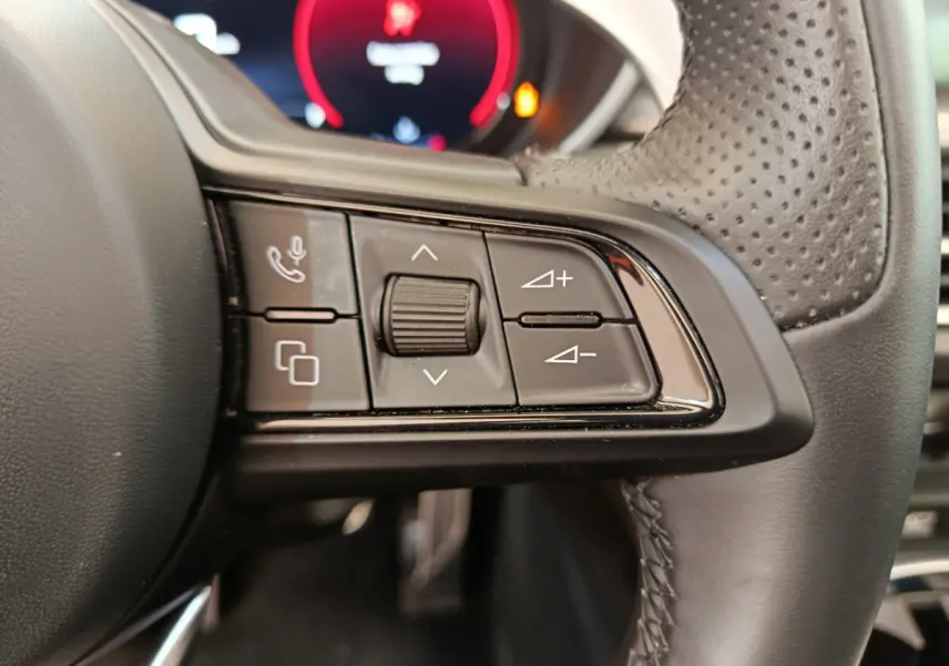 Gros plan sur les commandes gauche du volant en cuir perforé de l'Alfa Romeo Stelvio gris foncé, avec affichage numérique rouge en arrière-plan.