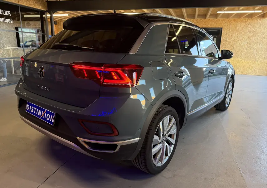 Vue 3/4 arrière droite d’un Volkswagen T-Roc gris métallisé 2022 avec feux arrière allumés, en intérieur showroom.
