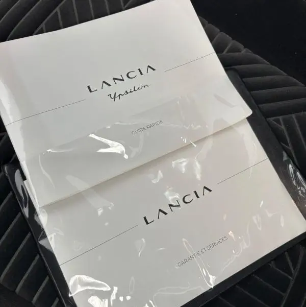 Image montrant deux manuels Lancia Ypsilon posés sur un siège en tissu gris foncé à motifs géométriques, sans vue du véhicule.