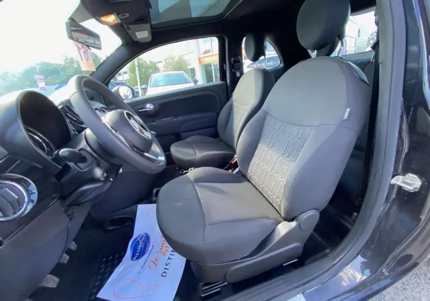 Intérieur côté conducteur d'une FIAT 500 noire 2024, sièges noirs avec motif FIAT, volant et toit panoramique visibles.