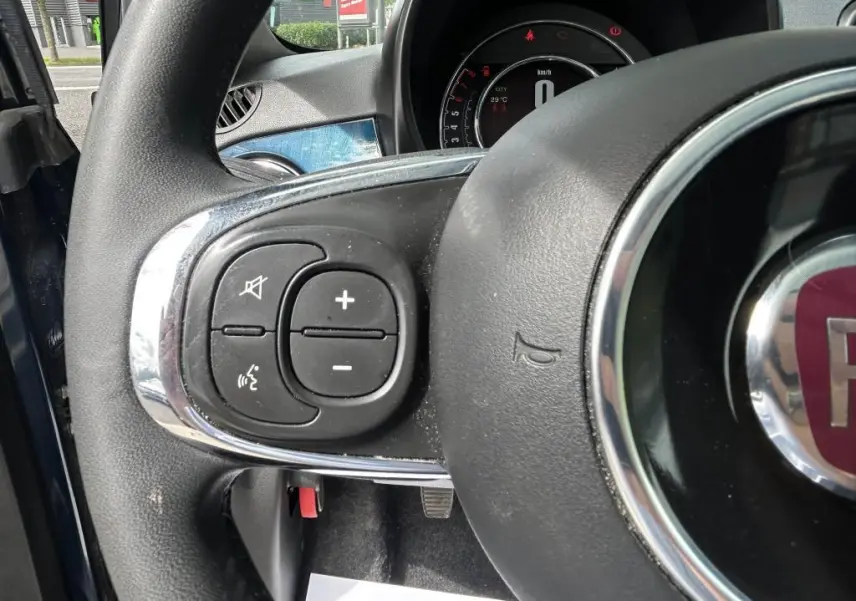 Gros plan sur les commandes au volant gauche du Fiat 500 bleu 2024 avec tableau de bord numérique en arrière-plan.