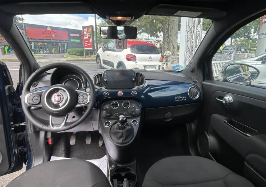 Vue intérieure avant du Fiat 500 bleu 2024 avec tableau de bord brillant, volant multifonction et levier de vitesses manuel.