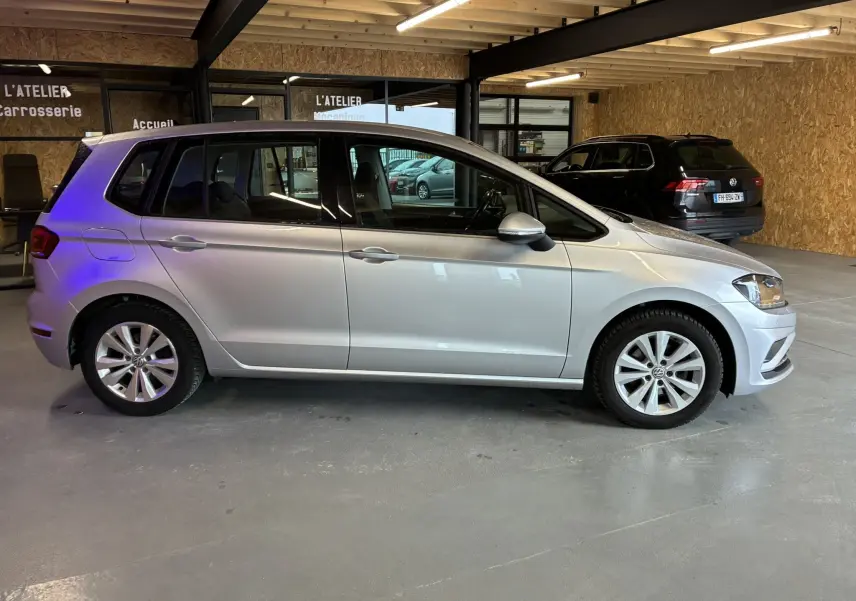 Volkswagen Golf Sportsvan gris vue de profil côté gauche dans un garage avec un autre véhicule noir en arrière-plan.