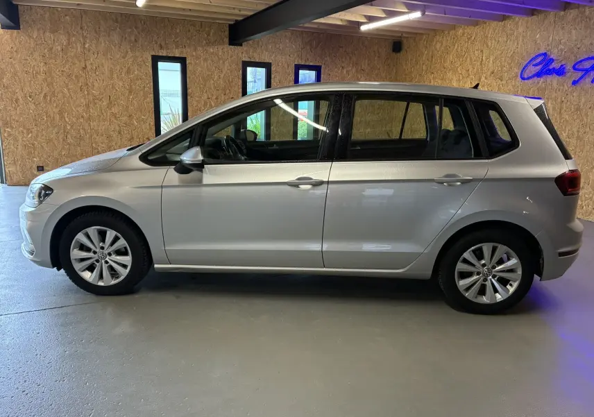 Profil côté gauche d'une Volkswagen Golf Sportsvan gris clair, 5 portes, dans un intérieur avec mur en bois.