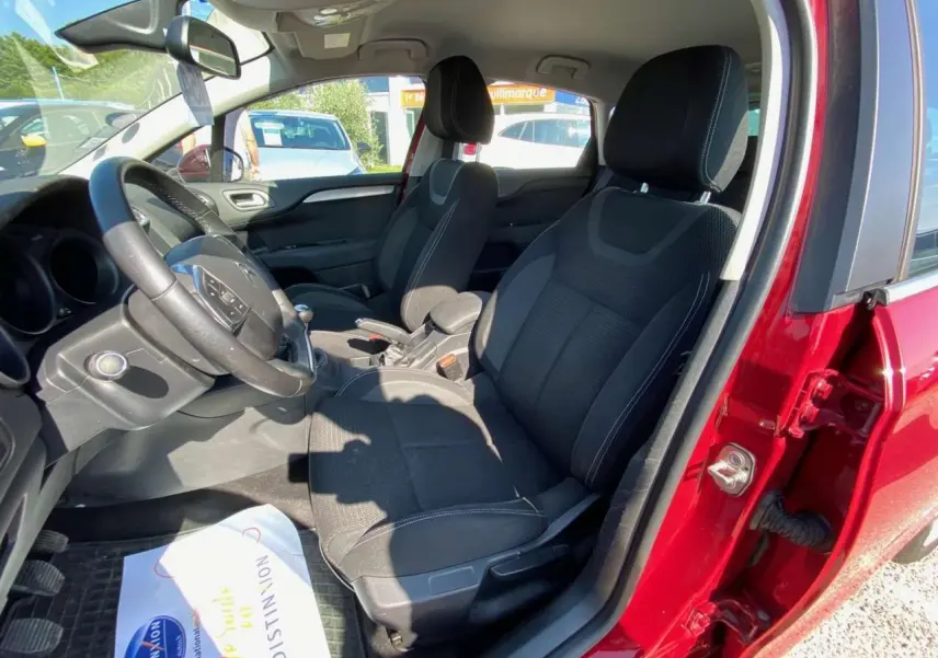 Vue intérieure côté conducteur d'une Citroën C4 rouge 2015, sièges noirs et volant avec commandes intégrées.