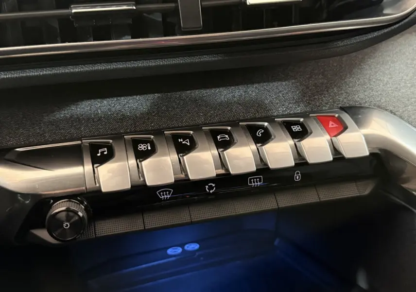 Gros plan sur la rangée de boutons argentés et noirs du tableau de bord du Peugeot 5008, éclairage bleu en dessous.
