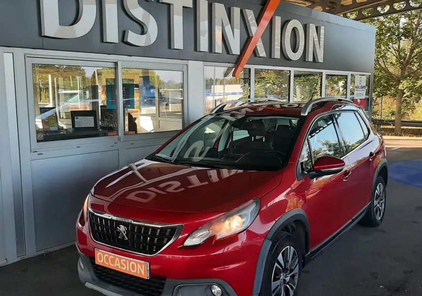 Peugeot 2008 rouge vue 3/4 avant droit, avec feux allumés et toit ouvrant, stationnée devant un panneau Distinxion.