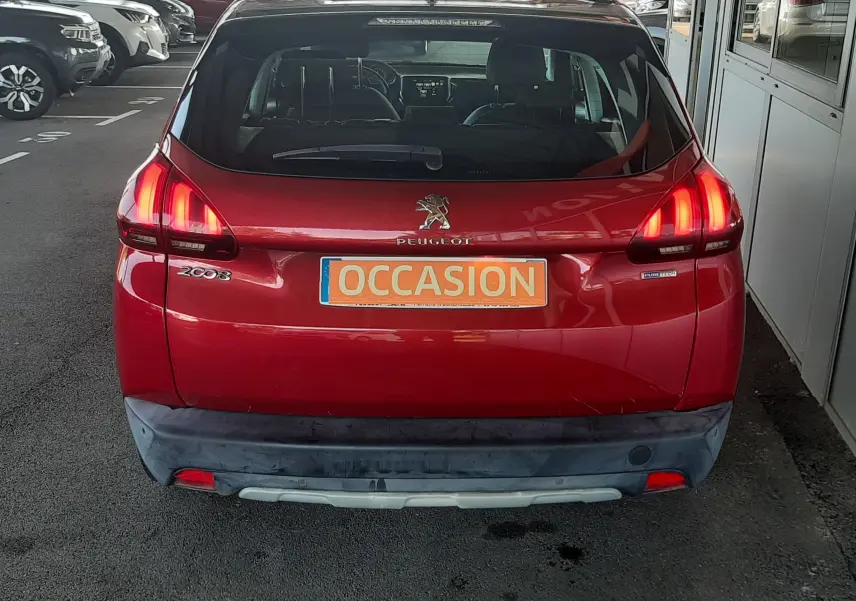 Vue arrière d'un Peugeot 2008 rouge avec feux allumés et plaque "OCCASION" sur un parking extérieur.