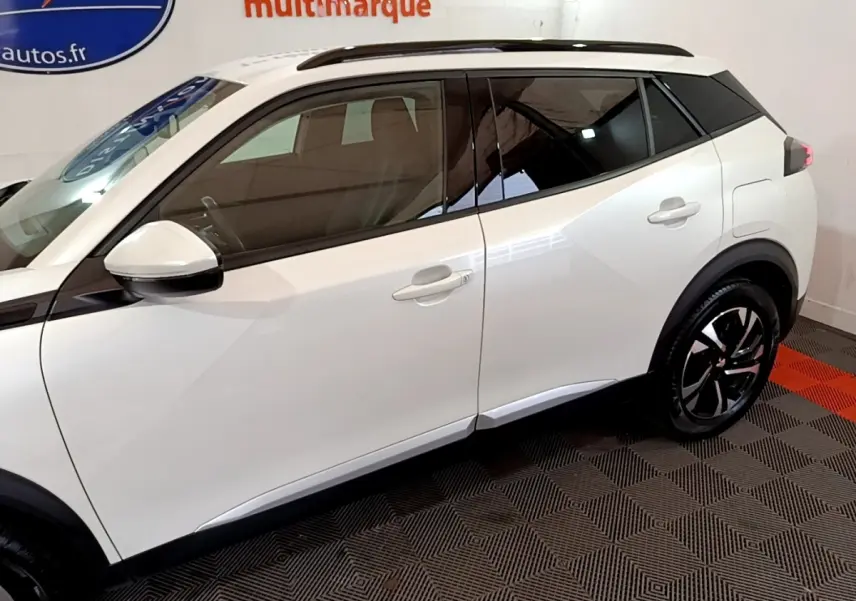 Vue latérale droite d’un Peugeot 2008 blanc avec toit noir et jantes alliage distinctives en intérieur showroom.