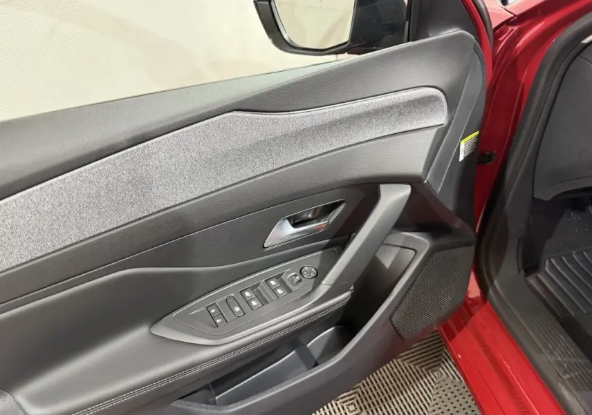 Vue intérieure sur la porte avant gauche rouge d'une Peugeot 308 2025 avec commandes électriques et habillage tissu gris.