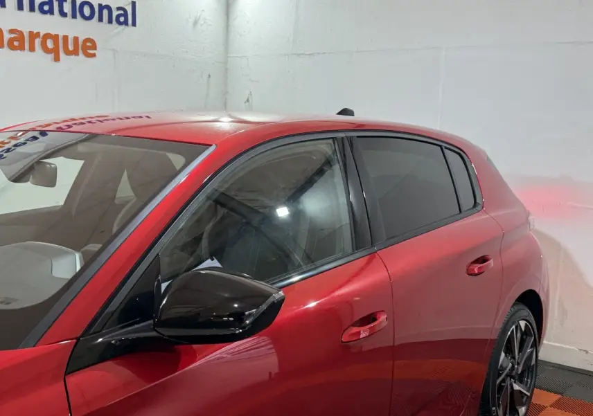 Vue côté gauche avant d'une Peugeot 308 rouge 2025 avec rétroviseur noir brillant et vitres teintées.