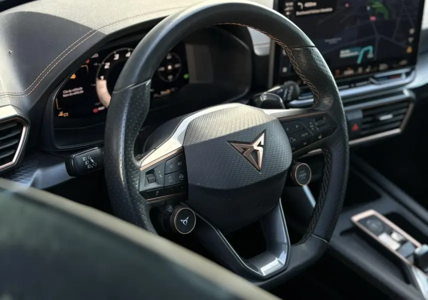 Vue rapprochée du volant cuir perforé noir avec logo CUPRA cuivre et commandes intégrées, tableau de bord numérique en arrière-plan.