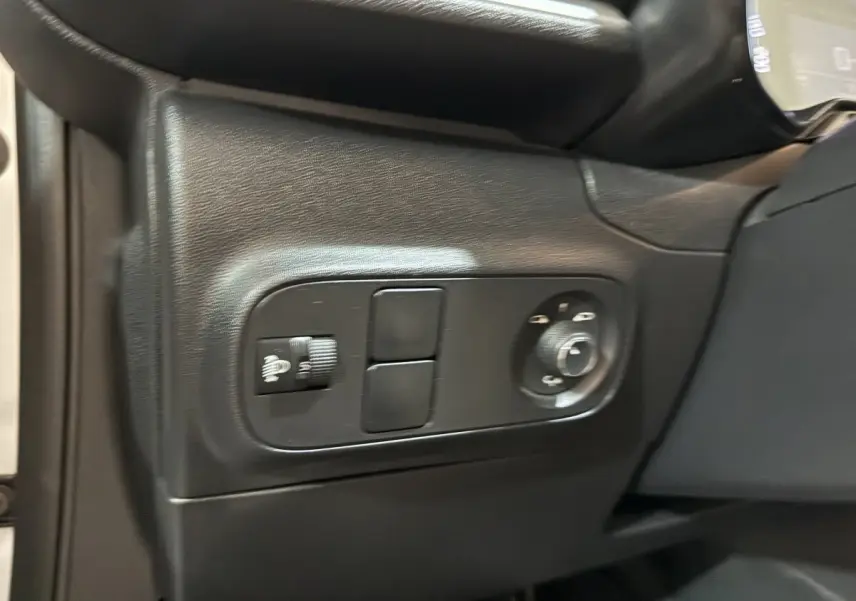 Gros plan sur les commandes de réglage des phares et rétroviseurs côté gauche de la planche de bord noire d'une Citroën C3 blanche.