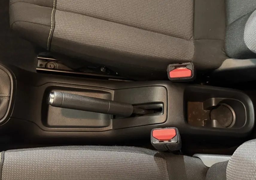 Vue rapprochée de la console centrale avec frein à main manuel et ceintures rouges entre les sièges avant tissu gris.