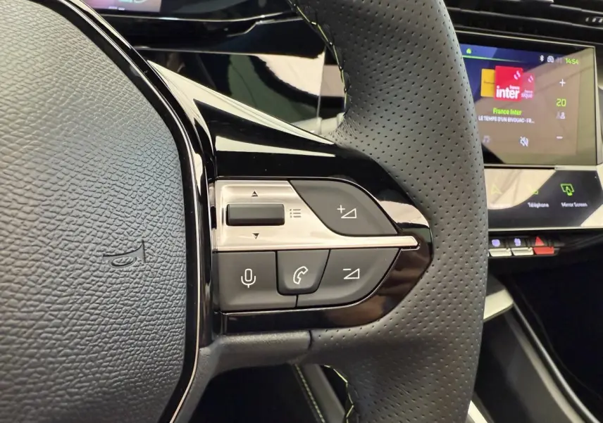Gros plan sur le volant cuir perforé gris foncé de la Peugeot 308 Hybrid 2024 avec commandes vocales et écran tactile en arrière-plan.