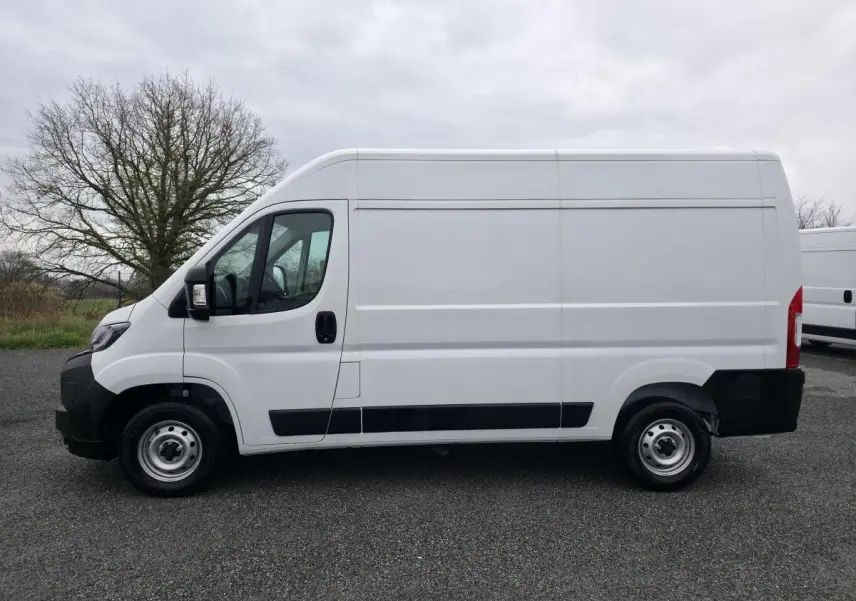 Peugeot Boxer Fourgon blanc en vue de profil côté gauche sur un sol goudronné, fond d'arbre et ciel nuageux.