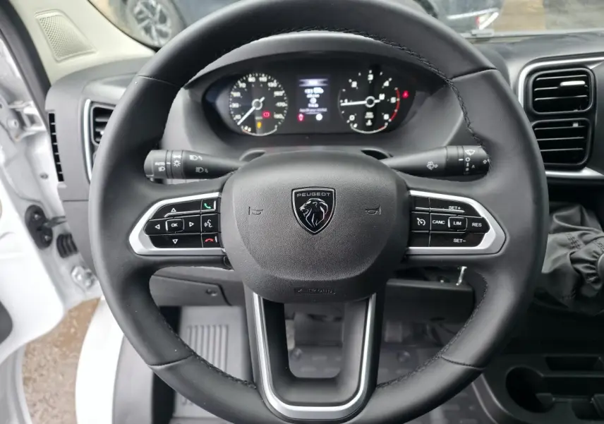 Vue rapprochée du volant cuir noir avec commandes intégrées du Peugeot Boxer Fourgon blanc, tableau de bord et compteurs en arrière-plan.
