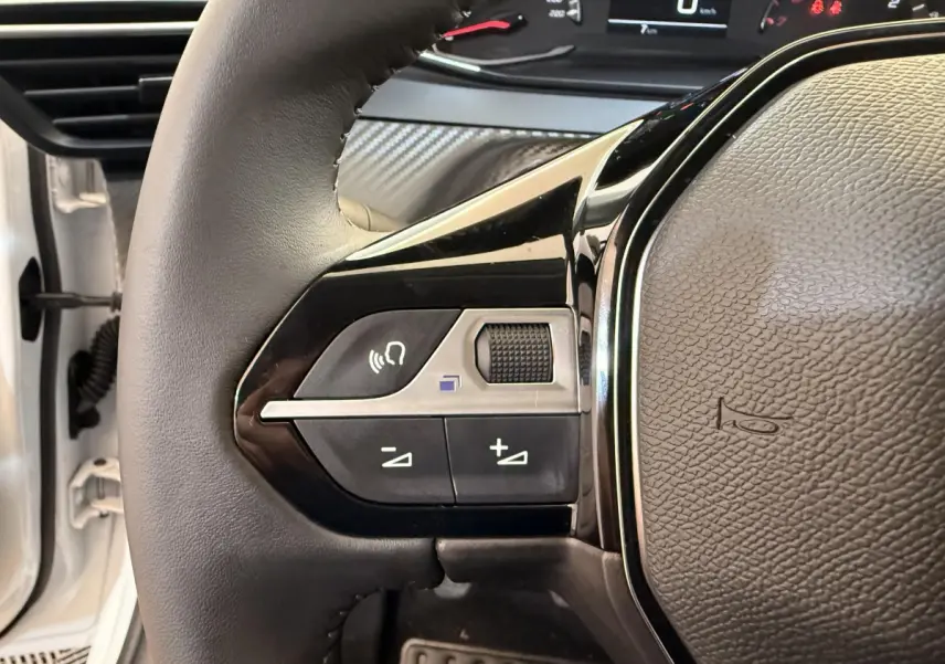 Gros plan sur le volant cuir noir de la Peugeot 208 blanche, avec commandes audio intégrées et tableau de bord partiel visible.