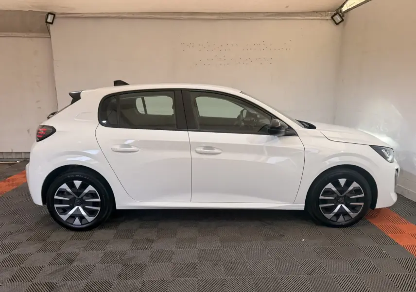 Profil droit d'une Peugeot 208 blanche 2025 avec jantes alliage et rétroviseurs noirs dans un garage.