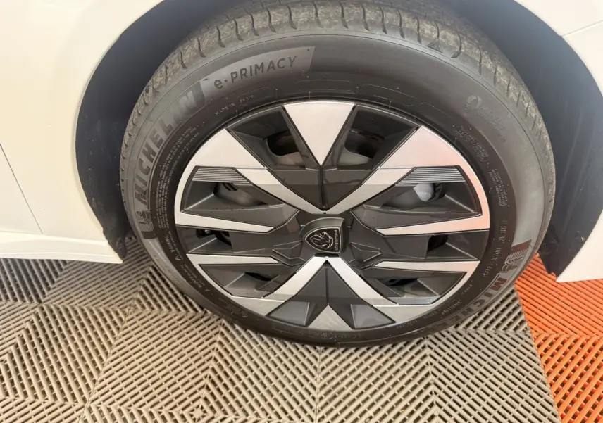 Gros plan sur la roue avant gauche d'une Peugeot 208 blanche, avec jante au design géométrique noir et argent.
