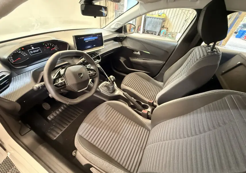 Vue intérieure côté conducteur de la Peugeot 208 blanche, montrant le tableau de bord moderne et les sièges gris à rayures.