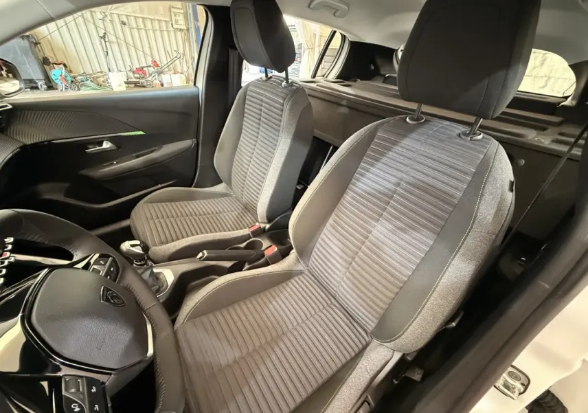 Vue intérieure côté conducteur de la Peugeot 208 blanche 2025, montrant les sièges avant en tissu gris et volant cuir.