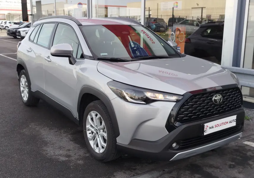 Toyota Corolla Cross gris acier vue 3/4 avant droit, avec calandre noire et phares allumés, stationnée devant un garage.