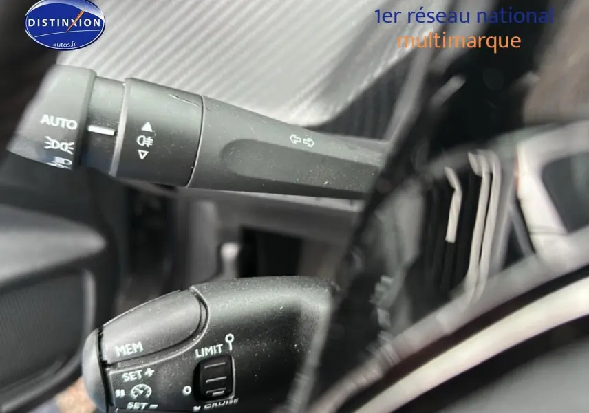 Détail du commodo gauche noir avec commandes de phares et régulateur sur Peugeot 208 gris Artense, intérieur proche.