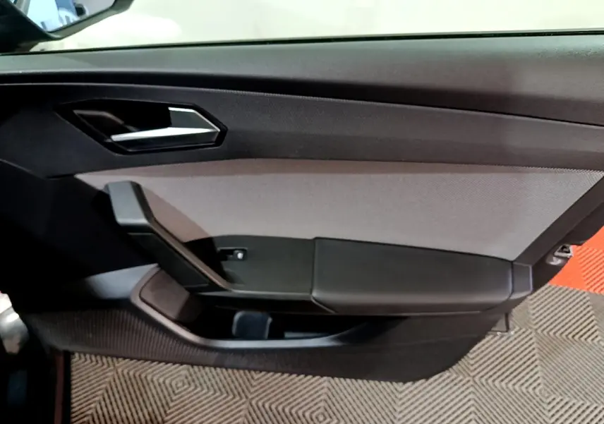 Vue rapprochée de la porte intérieure côté gauche de la SEAT Leon gris clair, avec poignée argentée et panneau noir texturé.