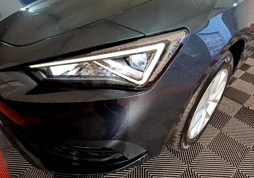 Gros plan sur le phare avant droit allumé d’une SEAT LEON gris clair, avec jante alu visible et carrosserie mouillée.