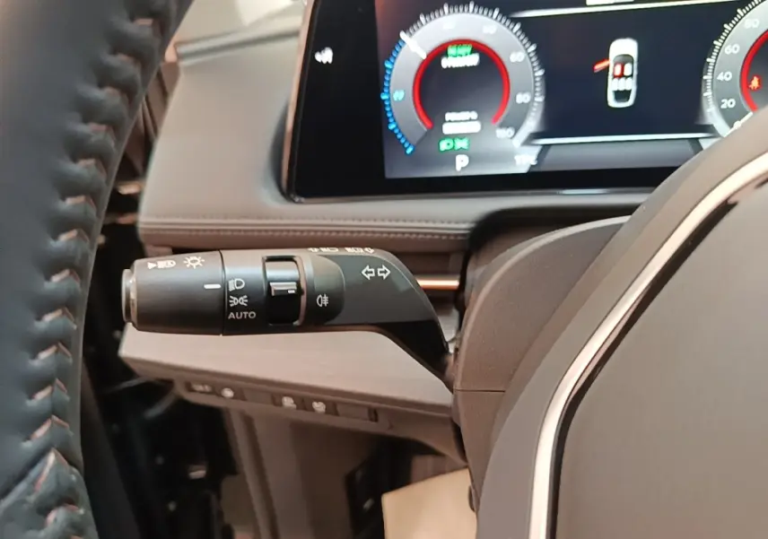 Gros plan sur la manette de clignotants et phares à gauche du volant cuir du Nissan Ariya noir, tableau de bord numérique visible.