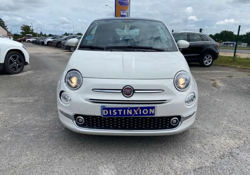 Vue de face d'une Fiat 500 blanche 2024 avec phares allumés et toit panoramique visible en arrière-plan.